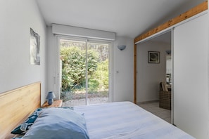 Une chambre à coucher comprenant un lit, une table de chevet, une lampe, une porte-fenêtre coulissante donnant sur un balcon et un panier en osier.