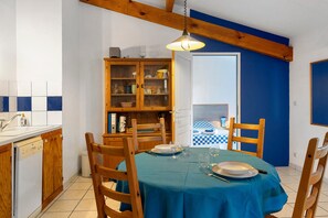 A dining area with a table set for four, wooden chairs, a cabinet with dishes, and a view of a bedroom with a checkered bedspread. - Apartment '4p Sanguinet-Doum Doum' with Outdoor Space (Sanguinet)
