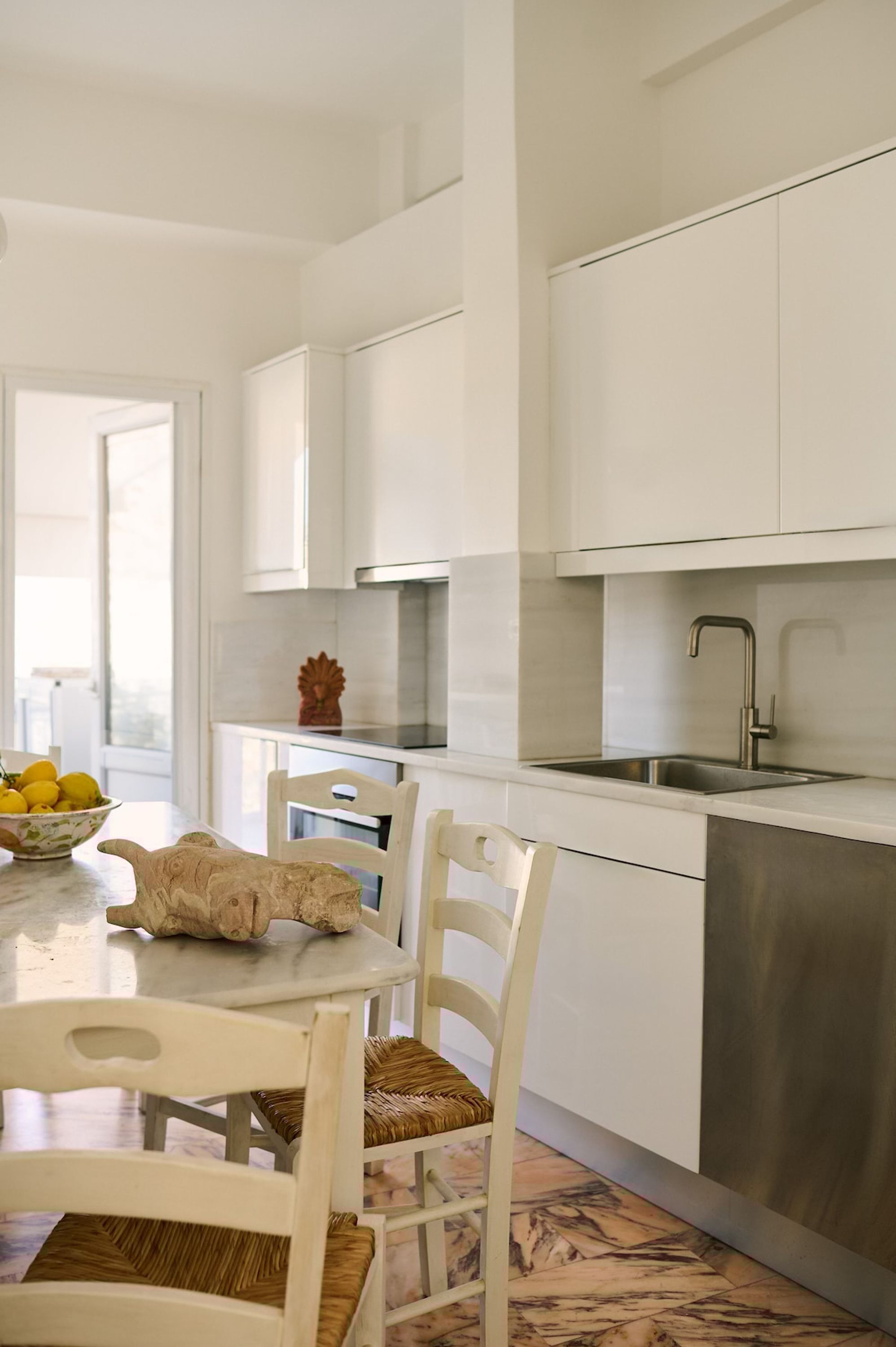 A modern kitchen with white cabinetry, a stainless steel sink, and a dining area with white chairs and a marble table.