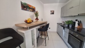 Una cucina compatta con un piano di lavoro in legno, una sedia nera, una ciotola di frutta, una pianta in vaso ed elettrodomestici in acciaio inossidabile.