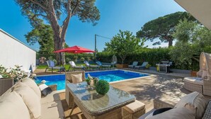 Ein Garten mit Schwimmbecken, Liegestühlen, einem Tisch mit Glasplatte und einem Grill.
