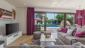 Ein modernes Wohnzimmer mit einem Flachbildfernseher, einem grauen Sofa, einem Couchtisch und einer großen Schiebeglasdoor, die zu einem Außenpoolbereich führt.
