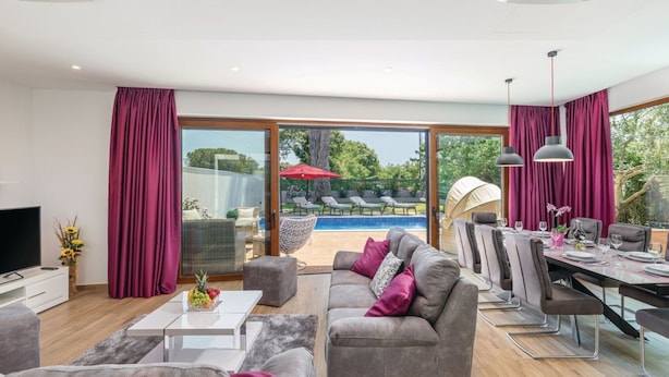 Ein modernes Wohnzimmer mit Blick auf einen Pool, grauen Sofas und einem Essbereich mit Tisch für vier Personen.