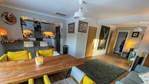 Ein modernes Wohnzimmer mit einem Holzesstisch, gelben Stühlen, einem grauen Sofa und einer Küche im Hintergrund.
