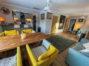 Ein modernes Wohnzimmer mit einem Holzesstisch, gelben Stühlen, einem grauen Sofa und einer Küche im Hintergrund.