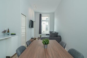 A modern living room with a wooden dining table, grey sofa, and a TV mounted on the wall.