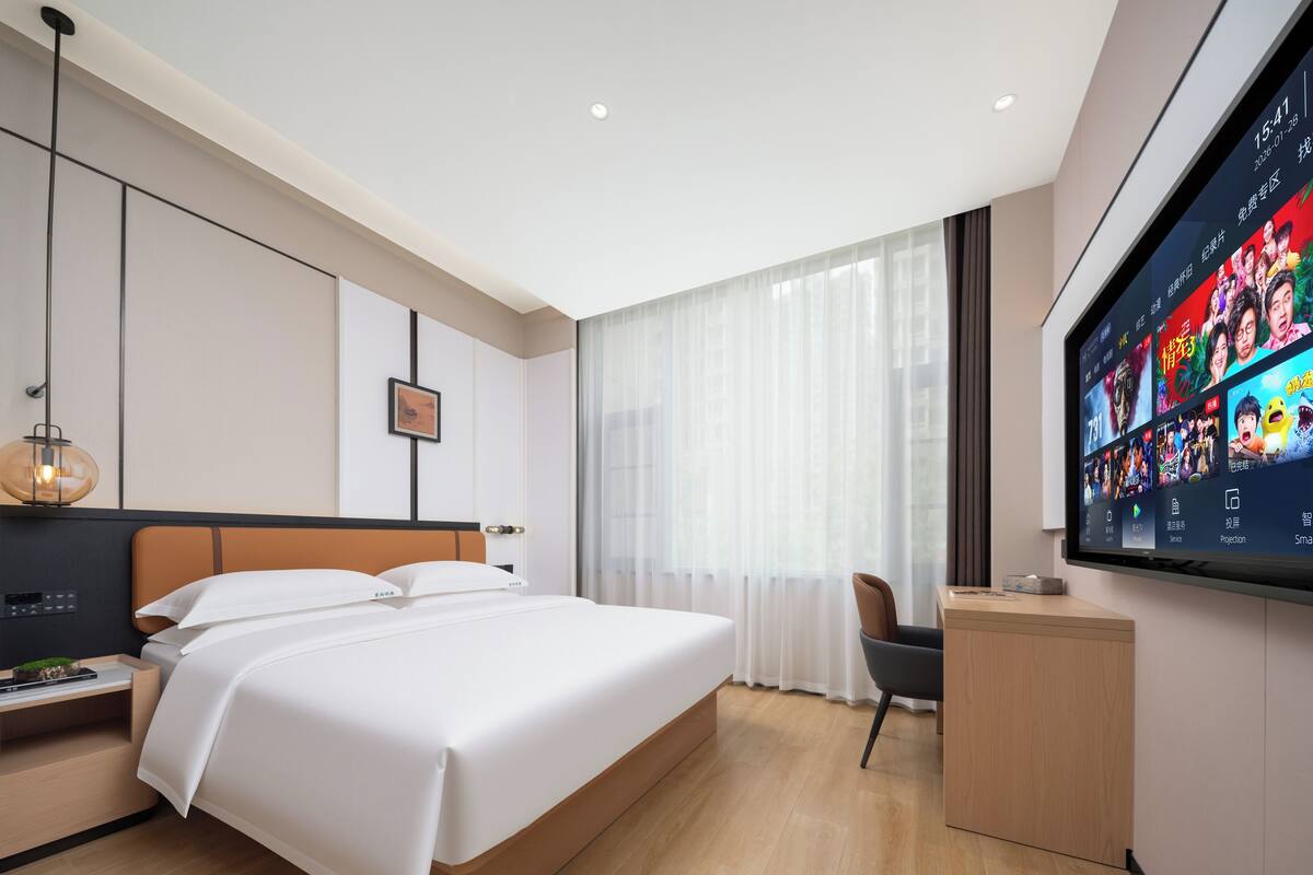 A modern hotel room with a large bed, a desk, a flat-screen TV, and a large window with sheer curtains.