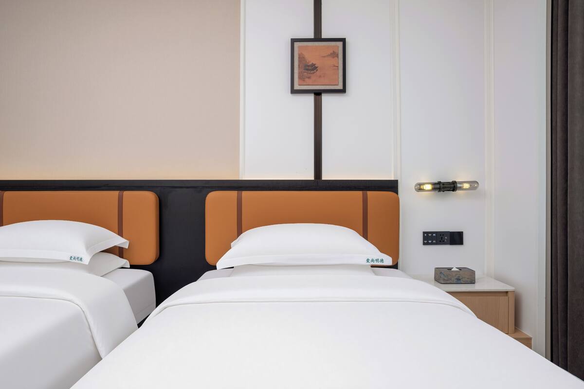 A hotel room with two beds, a nightstand with a lamp, a framed picture on the wall, and a power outlet.