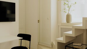 A minimalist room with a black chair, a flat-screen TV, a white vase with a plant, and a staircase. - N46 Studio Apartment  (Warsaw)