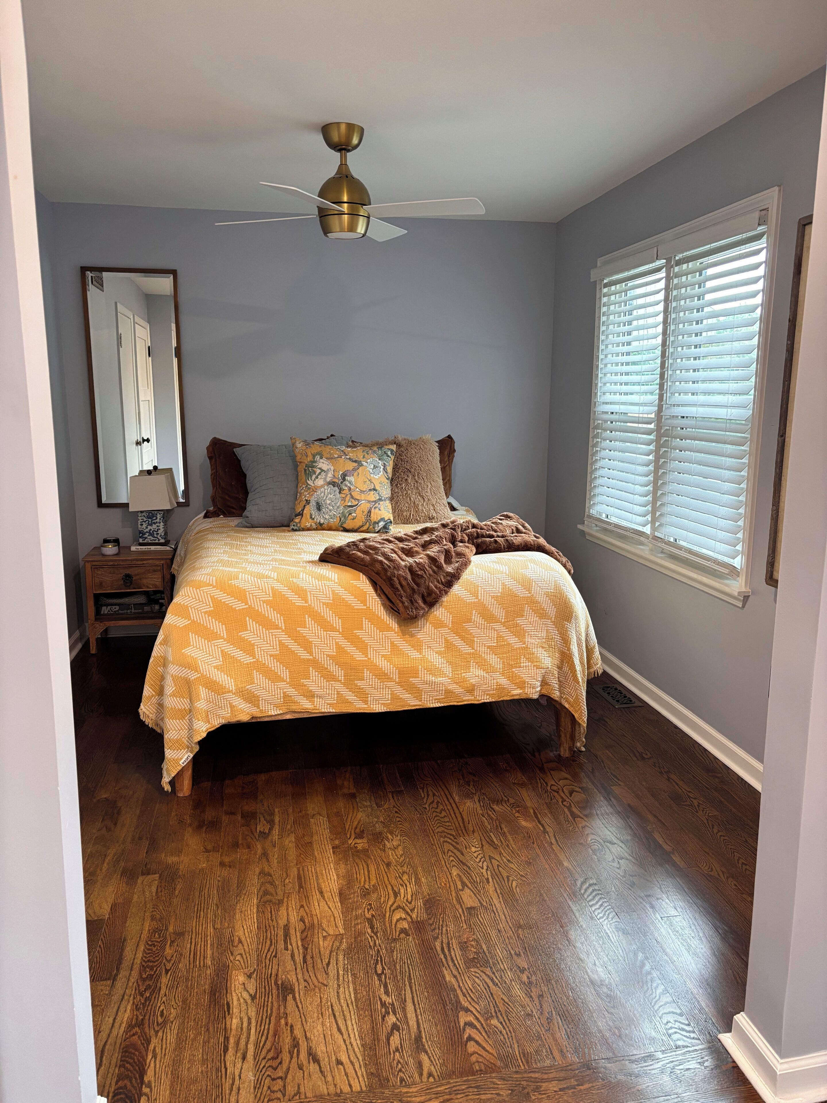 A bedroom with a bed, a ceiling fan, a nightstand, a window with blinds, and a wooden floor.