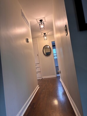 A hallway with wooden flooring, white walls, a door with a white doorbell, and a wall-mounted light fixture. - Eclectic, custom home! Central to all KC (Shawnee)