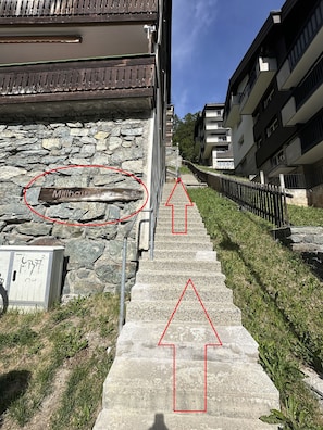 A stone wall with a wooden plaque reading 'Milhaus B.' and a concrete staircase leading to a building with balconies. - Matterhorn Alpine Hideaway - Cozy Renovated Apartm (Zermatt)