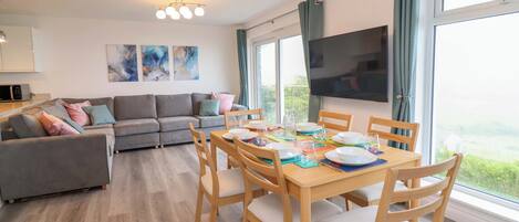 A modern living room with a grey sofa, wooden dining table, and chairs. There is a flat-screen TV mounted on the wall and a large window with curtains.