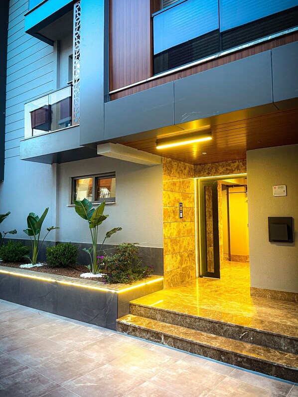 A modern building with a well-lit entrance, a small balcony, and a tiled pathway leading to the entrance. - Diker Port Residence (9) Mobilyalı ve Denize 5 Dk (konyaaltı)