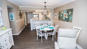 A dining area with a white table and chairs, a kitchen in the background, and artwork on the walls. - Beach Time, Fun Time, All the Time B505 at the Dunes! Fantastic View (Panama City Beach)
