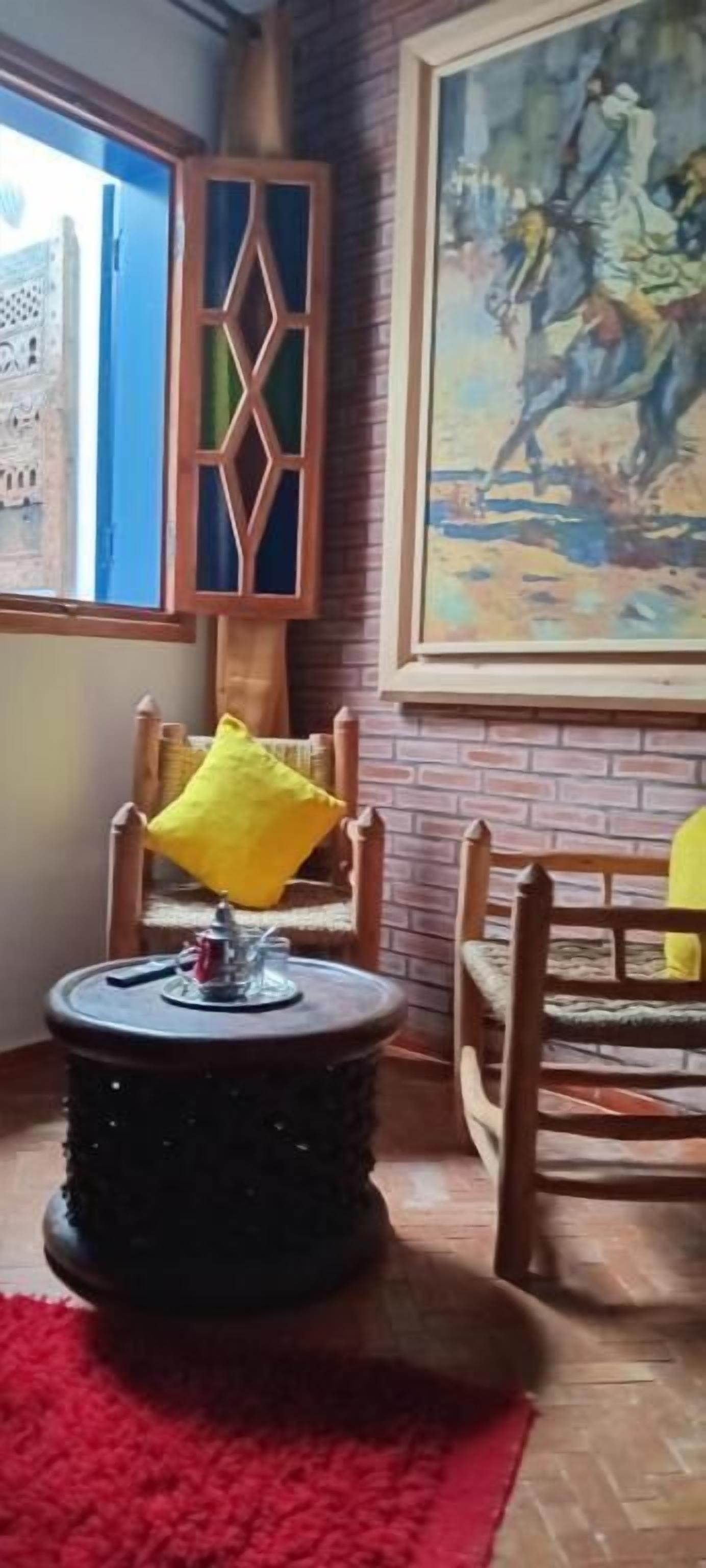 Um quarto com tapete vermelho, cadeiras de madeira, uma mesa redonda com almofada amarela, uma janela com vista para o azul e um quadro grande na parede.