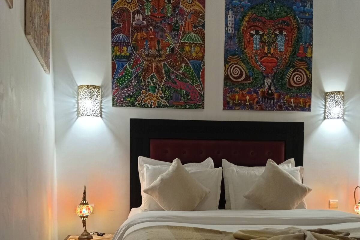 Una cama bien tendida con sábanas blancas, un cabecero rojo y dos lámparas fijadas a la pared.