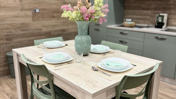 Een eethoek met een houten tafel, groene stoelen, een vaas met bloemen en een keuken op de achtergrond.