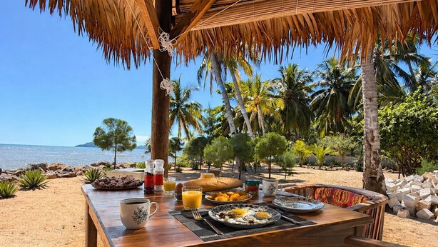 Una mesa de madera con comida para el desayuno, vista al mar y una estructura de techo de paja.