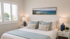 Une chambre à coucher comprenant un lit, deux tables de chevet, une commode, une lampe, un tableau et une fenêtre avec des stores.
