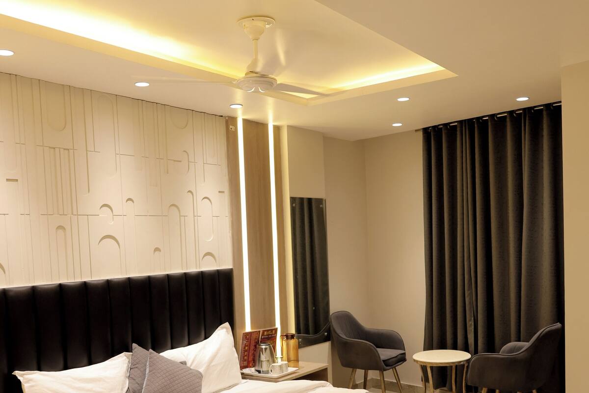 Habitación de hotel moderna con una cama grande, ventilador de techo, una mesita con sillas y un ventanal con cortinas.