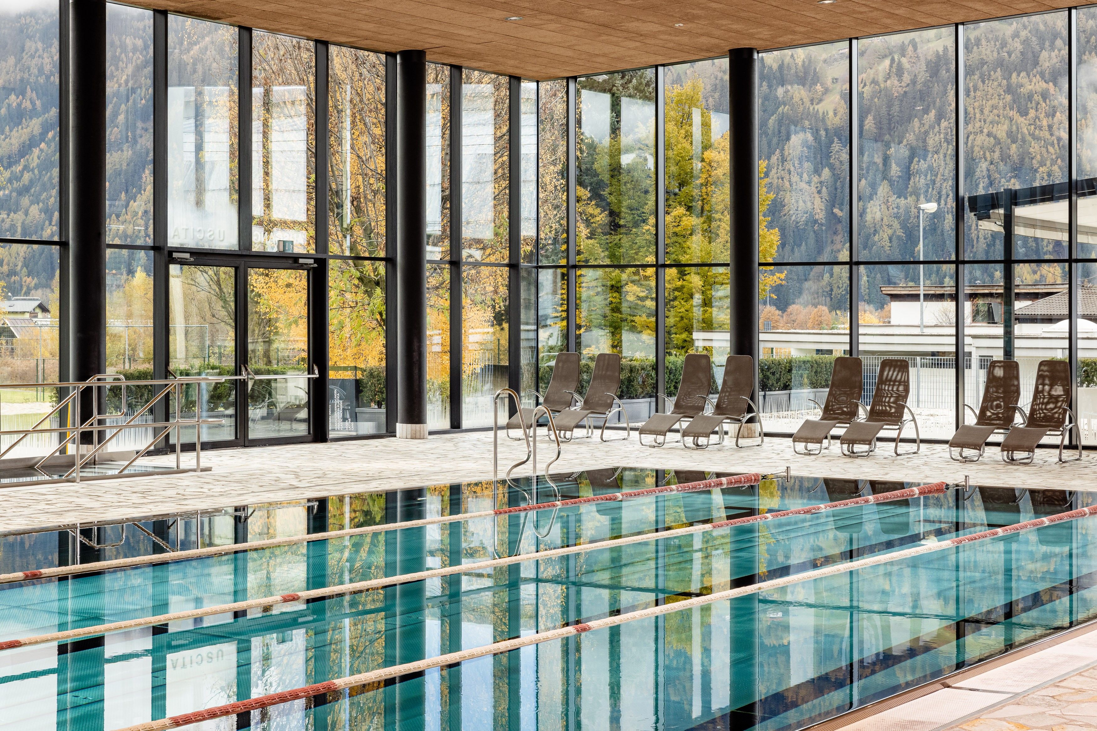 Una piscina con paredes de vidrio transparente, rodeada de sillones de descanso y vistas a un paisaje montañoso a través de amplios ventanales.