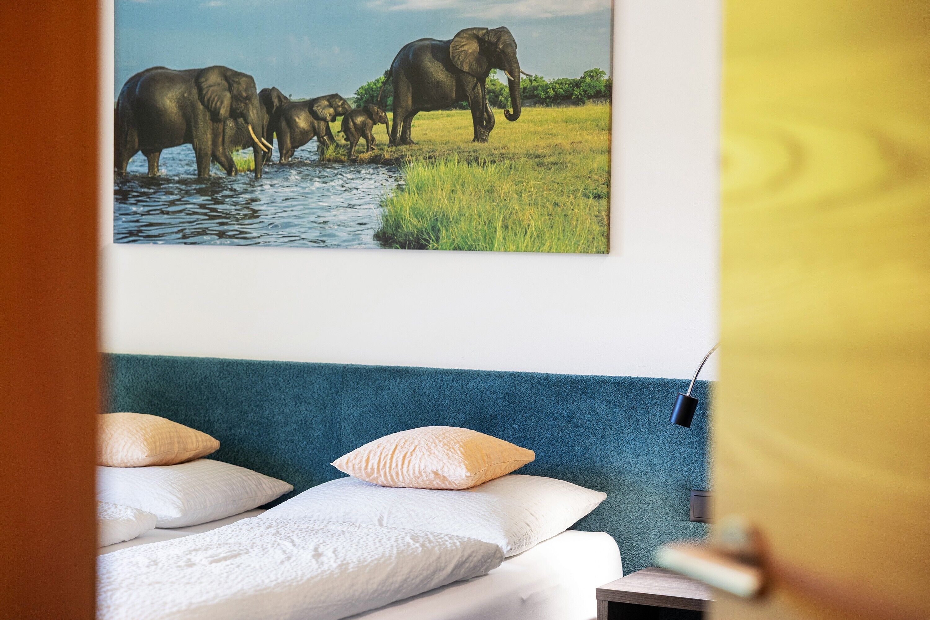 Een slaapkamer met een bed, kussens, een wandlamp en een schilderij van olifanten.