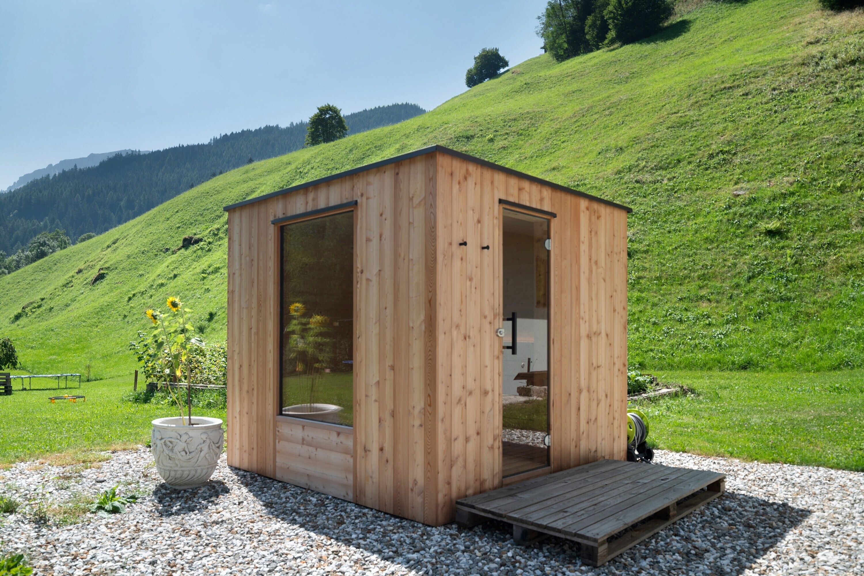 Una baita di legno con una porta a vetro, un piccolo patio e un giardino con un girasole.