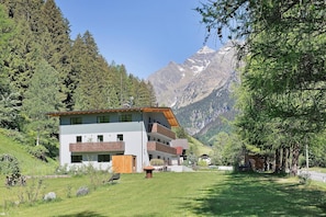 A modern multi-story building with balconies, surrounded by lush greenery and mountains in the background.