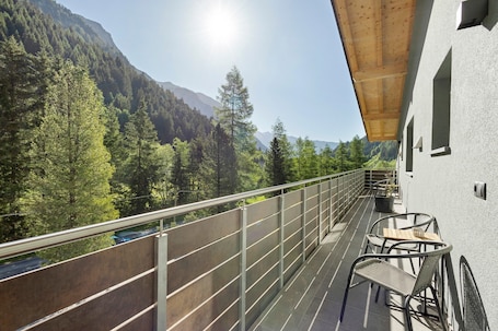 A balcony with a metal railing, a table, and two chairs, overlooking a forested mountain area.