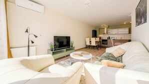 A modern living room with a flat-screen TV, a sofa, a coffee table, and a dining area with chairs.