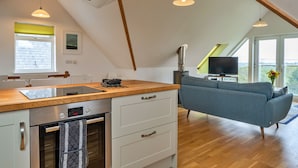 Et moderne køkken med indbygget ovn og træbordplade, en grå sofa og et fjernsyn i en opholdsstue med store vinduer.