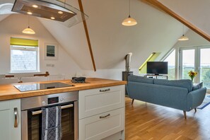 Eine moderne Küche mit Einbauofen und einer Holz Arbeitsplatte, ein graues Sofa und ein Fernseher in einem Wohnbereich mit großen Fenstern.
