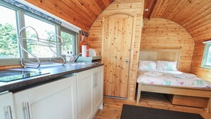 Ein gemütliches Holzhaus mit Küchenzeile, einem Bett mit bunter Bettdecke und einem Fenster mit Blick auf Grünflächen.