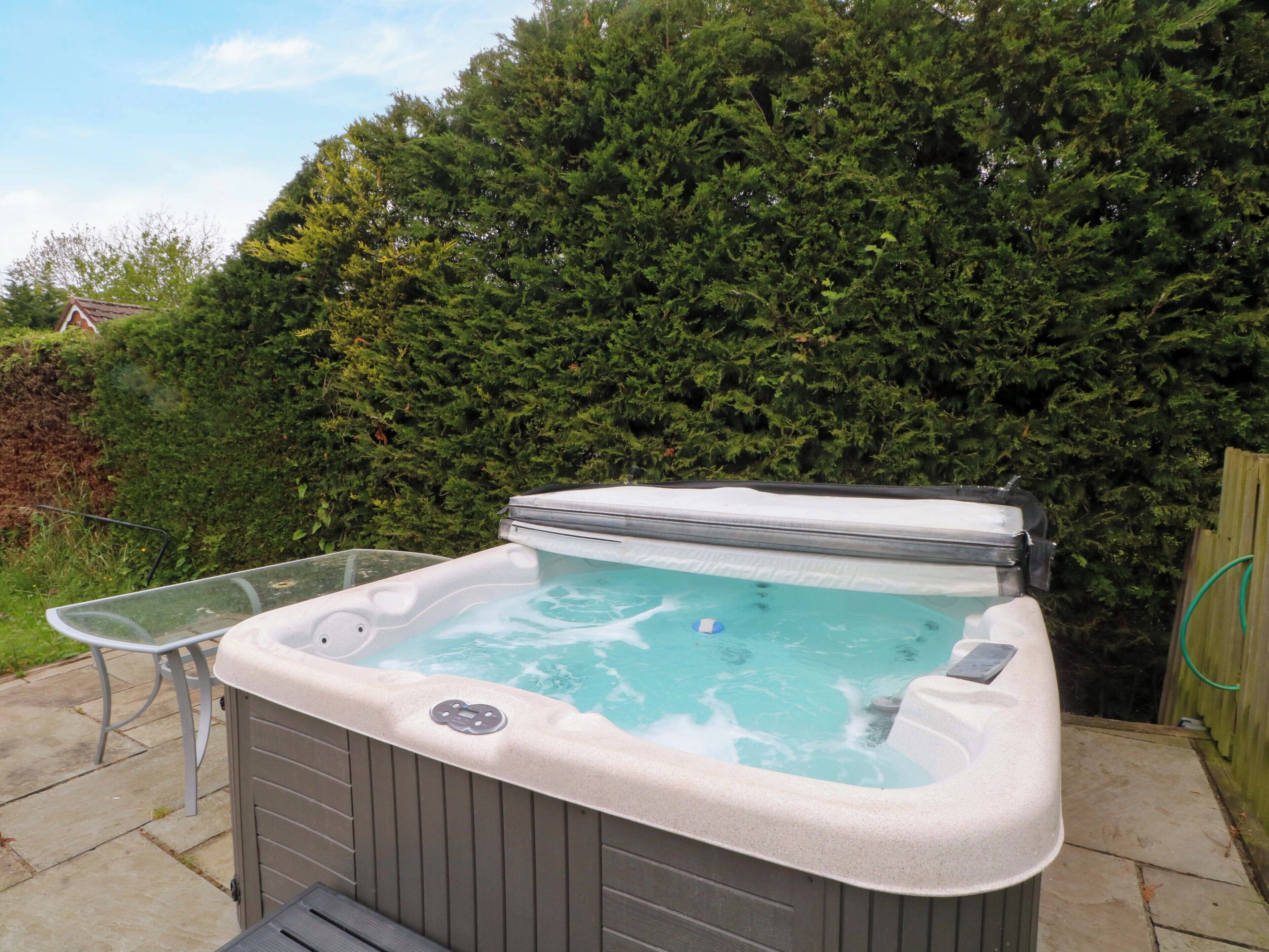 Un jacuzzi con una mesa y silla de vidrio, rodeado por un seto y una valla de madera.