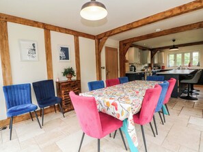Un comedor con una mesa larga, sillas coloridas, vigas de madera y una cocina al fondo.