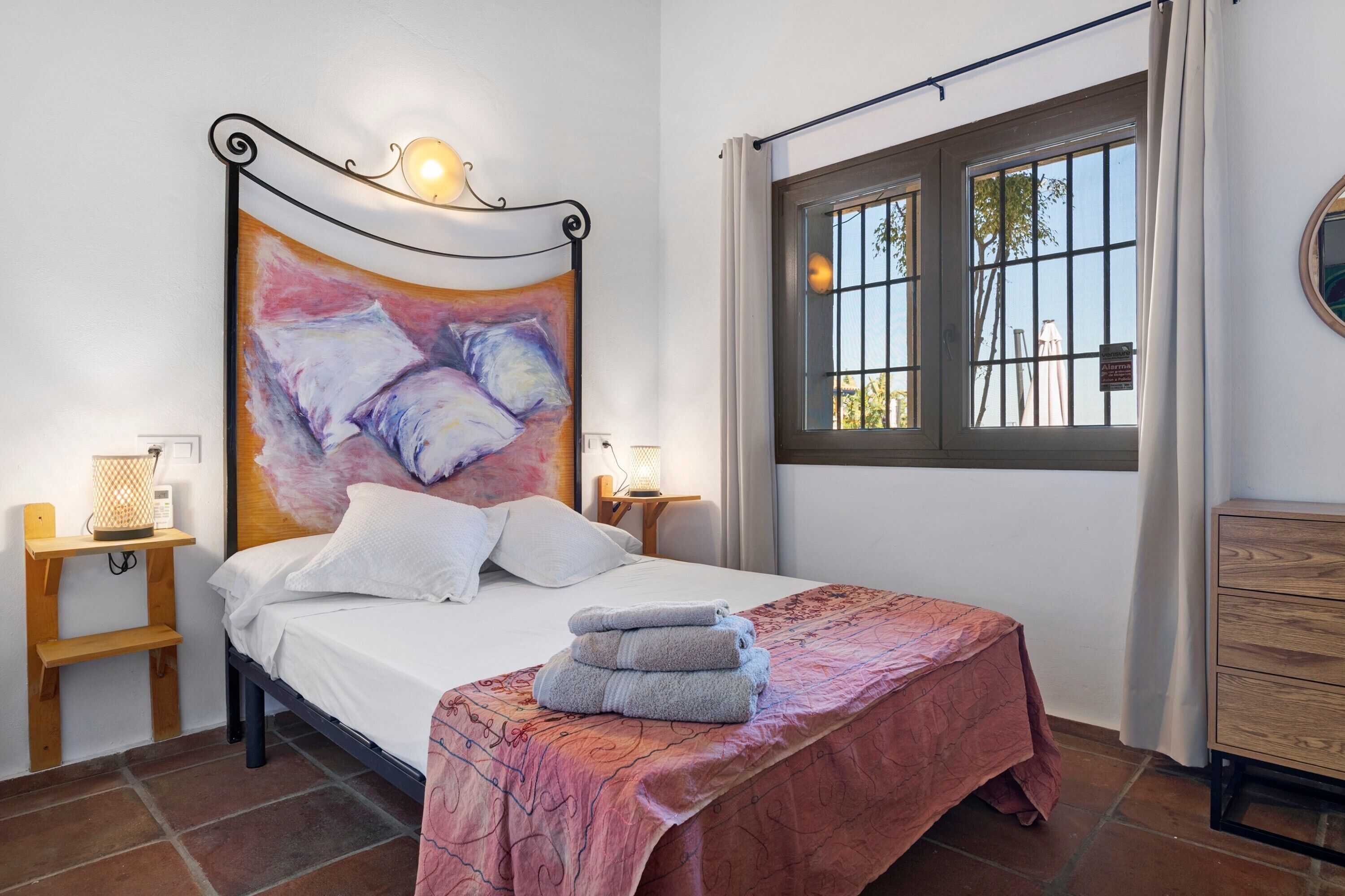 A bedroom with a bed, bedside lamps, a window with a view, a wooden dresser, and a painting on the wall.