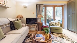 Una sala de estar con sofá, televisor, lámparas decorativas, una mesa de centro con libros y flores, y vistas a través de un ventanal.