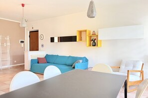 Uma sala de estar moderna com um sofá azul, cadeiras brancas, uma mesa de jantar e uma estante amarela com objetos decorativos.