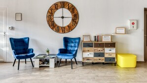 Ein Wohnzimmer mit zwei blauen Sesseln, einem Couchtisch, einer großen Wanduhr, einem Bücherregal und einem gelben Hocker.