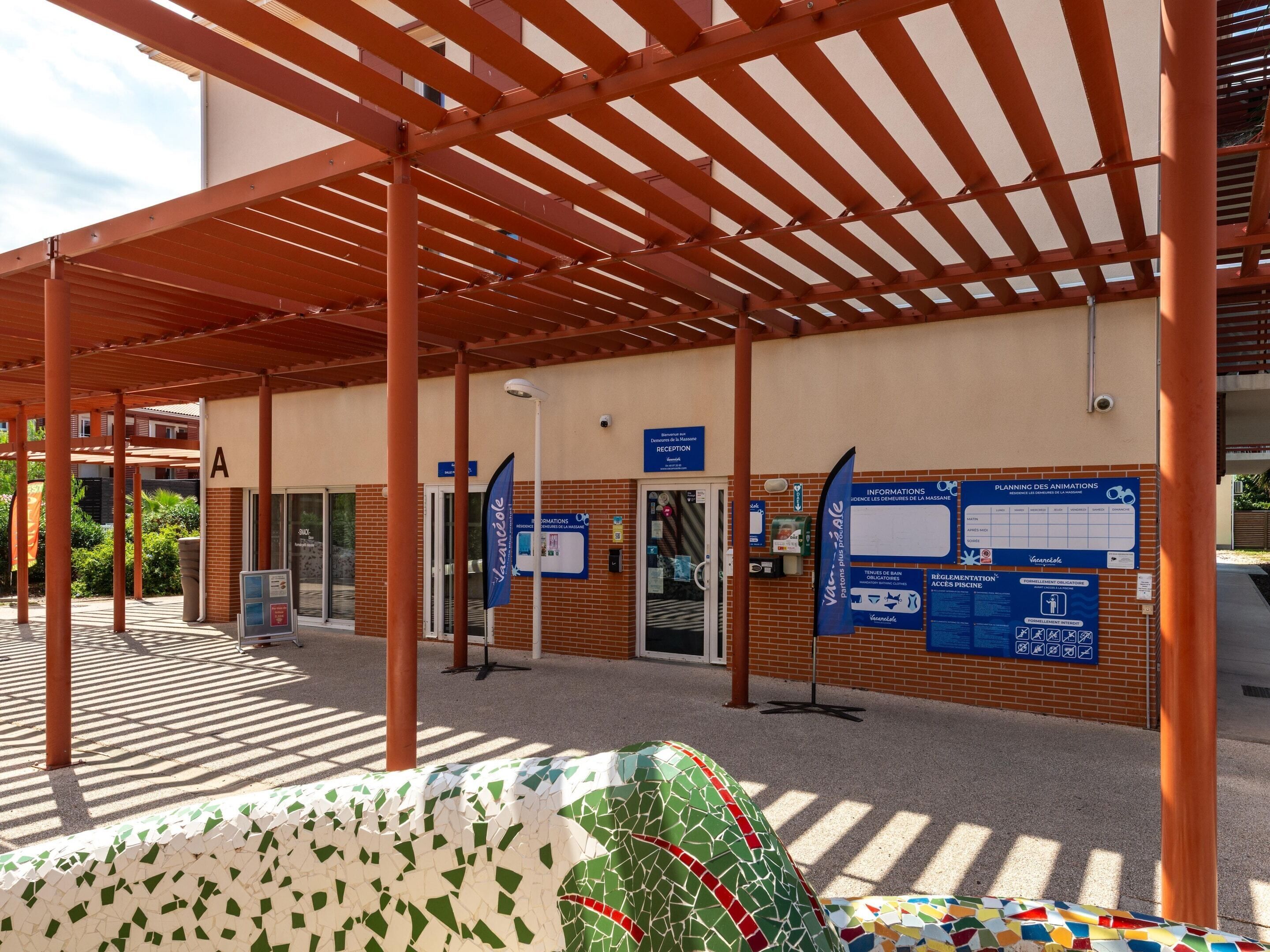 A covered outdoor area with a reception desk, informational signs, and a tiled floor.