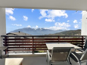 Ein Balkon mit Tisch und Stühlen, mit Blick auf ein Gebirge mit schneebedeckten Gipfeln und blauem Himmel.
