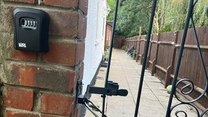 A metal gate with a padlock, a brick wall, and a white fence in the background. - House Sleeps 6 Garden (Mitcham)