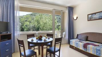 A room with a dining table, chairs, a sofa, a TV, and a window with a view of trees.
