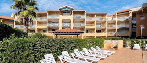 Un hotel con varios balcones, una zona de piscina con tumbonas y una palmera.