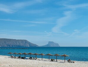 Ein Strand mit Sonnenschirmen und Liegestühlen, einem klaren Himmel und einem fernen Berg.