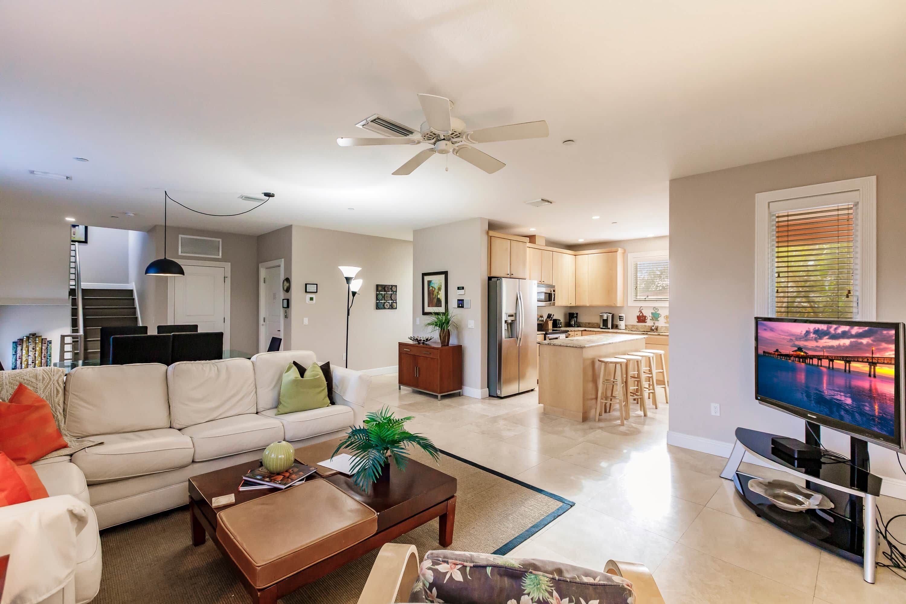 A modern living room with a sofa, coffee table, and a TV. There's a kitchen area with a refrigerator and a dining space.