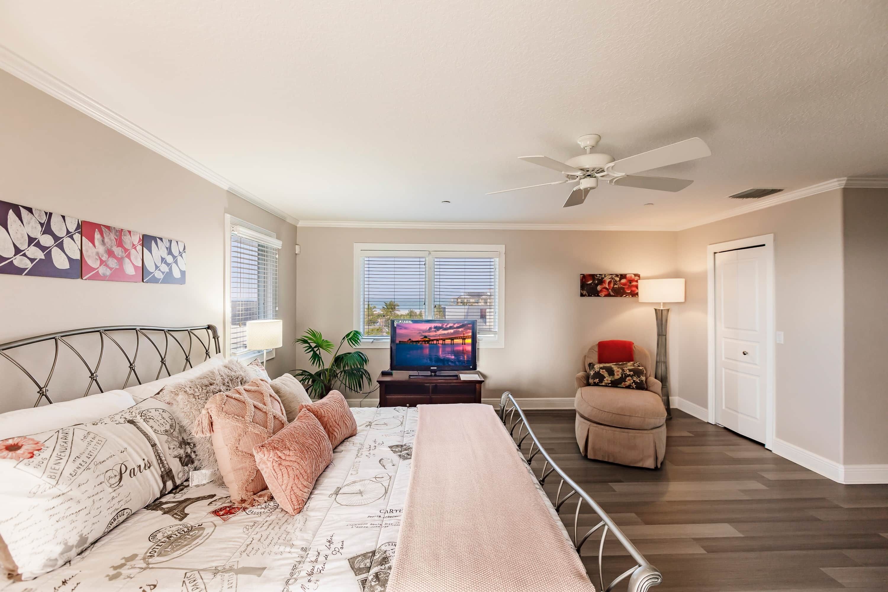 A bedroom with a bed, a TV, a ceiling fan, and a window with blinds.