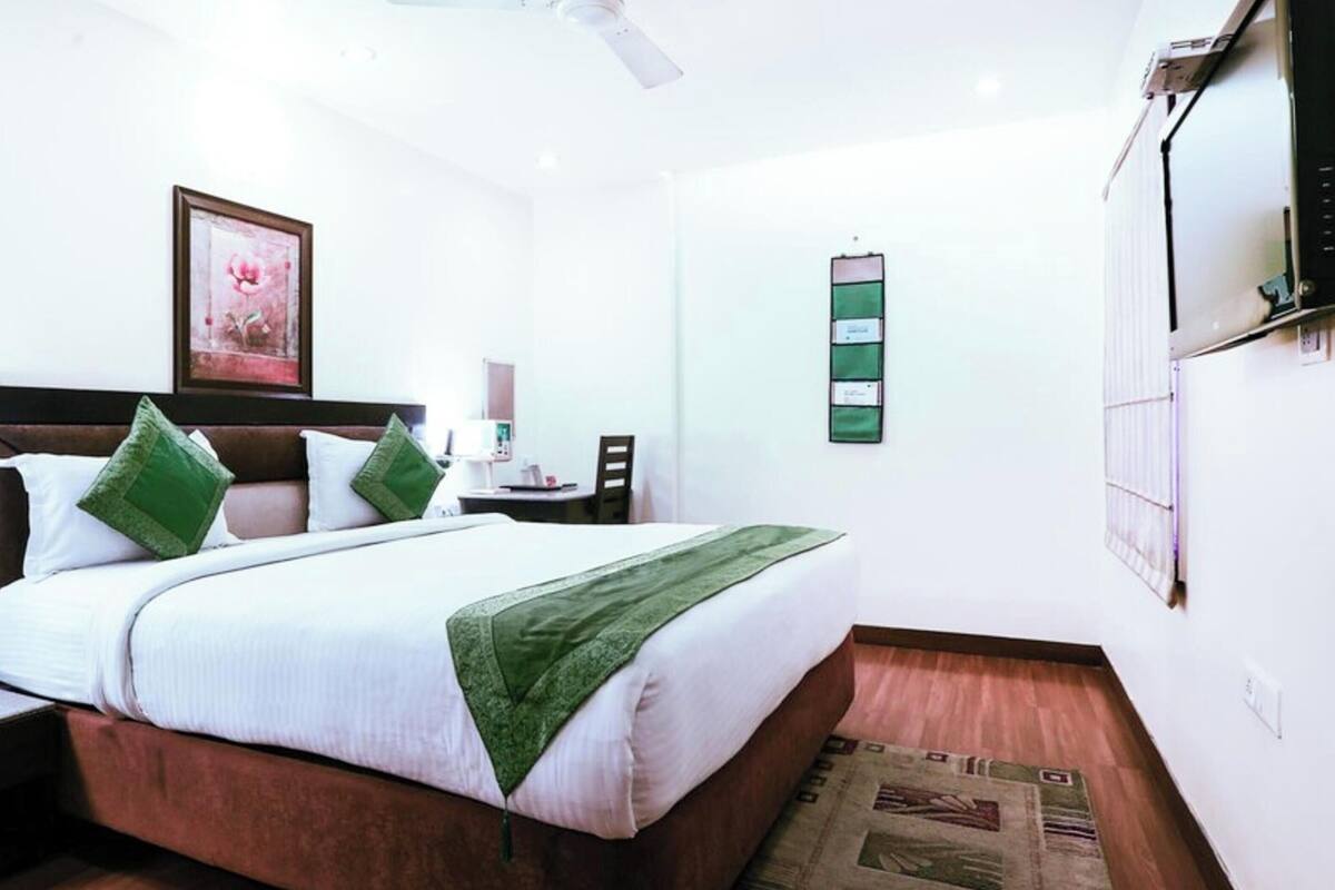 Quarto de hotel com cama, mesa de cabeceira, abajur e televisão presa à parede.
