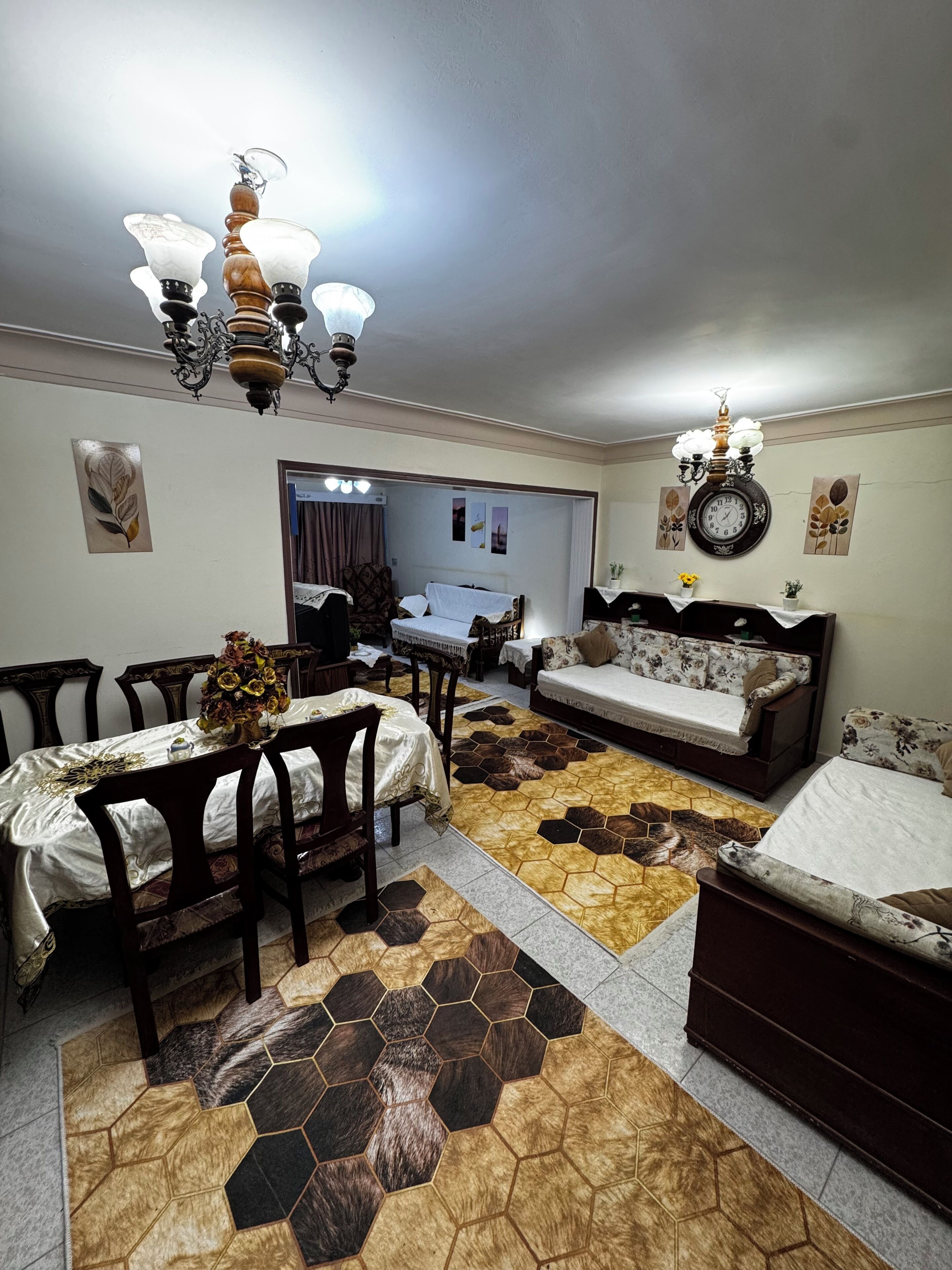 A room with a dining table, chairs, a sofa, a clock, and a chandelier.